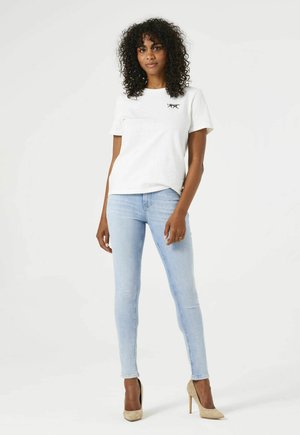 Weißes Baumwoll-T-Shirt mit einem kleinen schwarzen Grafik, kombiniert mit hellblauen engen Jeans und beigen High Heels. Das Model steht vor einem neutralen Hintergrund.