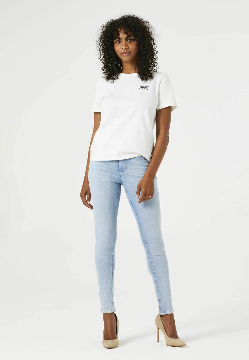 Camiseta blanca de algodón con un pequeño gráfico negro, combinada con jeans ajustados de color azul claro y zapatos de tacón beige. El modelo se encuentra en un fondo neutro.