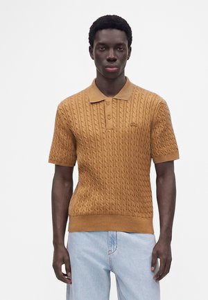 Jeune homme portant un polo marron à manches courtes en maille torsadée et un jean bleu clair, debout devant un fond blanc uni.