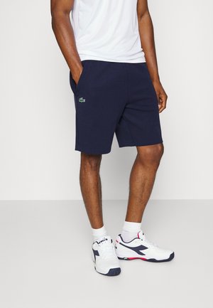 Man draagt marineblauwe korte broek met groen krokodillenlogo, wit T-shirt, witte sokken en witte sneakers met marineblauwe en rode accenten.