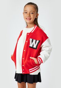 Rote und weiße Varsity-Jacke mit gerippten Akzenten, gesticktes "W" auf der Tasche, kombiniert mit einem weißen Pullover und einem schwarzen Rock.