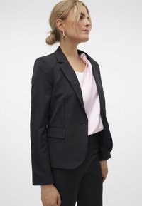 Svart skreddersydd blazer med revers, enkel knappelukking og to frontlommer, båret over en lys rosa topp med knytedesign.