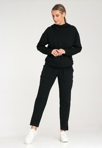 Czarna bluzka z kapturem i pasujące zwężane spodnie, wykonane z miękkiego materiału. Spodnie mają przednie kieszenie i rozcięcia przy kostce. Białe sneakersy.