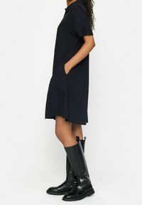 Abito blu navy a maniche corte con colletto e tasche laterali, caratterizzato da una vestibilità comoda e una texture liscia. Abbinato a stivali neri al ginocchio.
