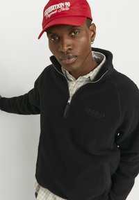 Sort fleece-pullover med lynlås ved kraven, der har en struktureret overflade og et præget logo. Båret med en ternet skjorte under.