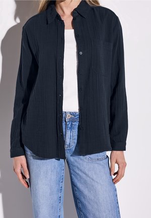 Vrouw draagt een losse zwarte blouse met knopen over een wit topje en lichtblauwe spijkerbroek, te zien van schouders tot heupen tegen een witte achtergrond.