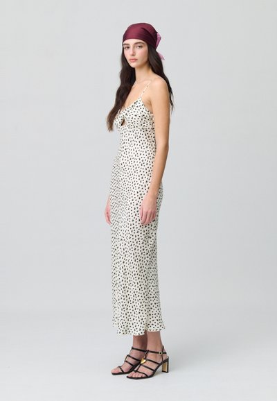 Vestido midi de lunares en crema con tirantes finos, escote en forma de V y un detalle con volantes. La modelo lleva unos tacones negros con tiras y un acento dorado.