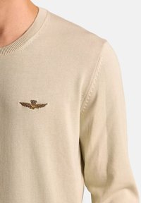 Maglione beige a maglia con scollatura rotonda. Presenta un piccolo logo ricamato con ali sul petto. Morbido al tatto e con un design aderente.