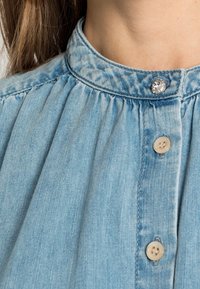 Ljusblå denimskjorta med en samlad krage, dekorativ klar knapp och klassisk knäppning med två beige knappar.