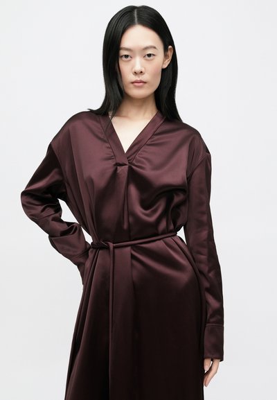 Μακρύ, βαθύ βυσσινί φόρεμα από σατέν. Διαθέτει V-λαιμόκοψη, μακριά μανίκια και ταιριαστή ζώνη δεμένη στη μέση. Λείο ύφασμα, μινιμαλιστικός σχεδιασμός.