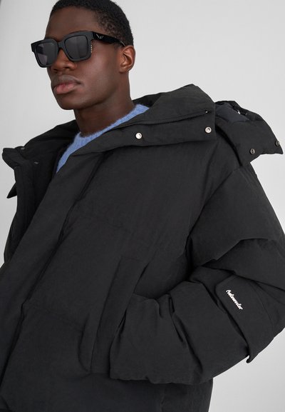 Black puffer jacket with a hood, featuring large quilted sections, zip closure, and a front pocket. Model wears black sunglasses and a blue sweater.