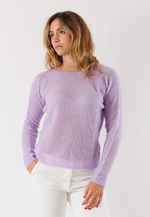 Donna con capelli biondi ondulati che indossa un maglione di lana lavanda e pantaloni bianchi, con lo sguardo di lato e le mani parzialmente in tasca.