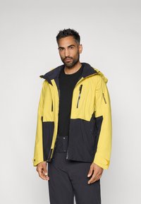 Geltonas ir juodas vandeniui atsparus džemperis su gobtuvu. Jame yra užtrauktukais užsegamos kišenės, smakro apsauga ir kontrastingos spalvos krūtinės ir rankovių srityse.