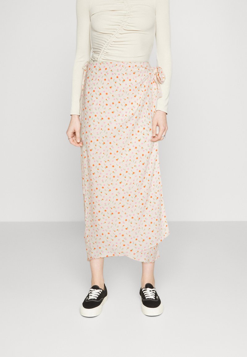 Blomsterindpakningsskørt i lys pink med små orange og pink blomster; letvægtsstof; har en sidebindedetajle; parret med sorte sneakers.