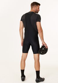 Svarta cykelbyxor i bib-stil gjorda av stretchy tyg, med vadderad sits, ryggsuspenders och en åtsittande design. En svart hjälm hålls i handen.