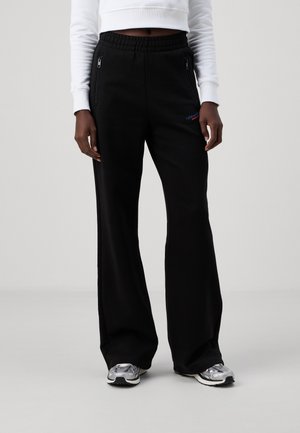 Osoba w czarnych szerokich dresach z kieszeniami na zamek, białej krótkiej bluzie oraz srebrnych tenisówkach, stojąca na tle jednolitego tła.