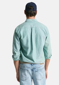 Lichtgroene langemouwen shirt met opgerolde mouwen, achterkant met plooi-detaillering, gedragen met lichtblauwe jeans. Donkere pet op het hoofd. Casual fit.