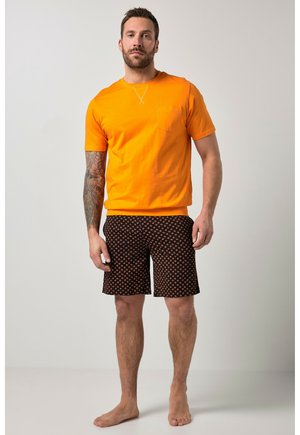 Mężczyzna stojący boso, ubrany w pomarańczową koszulkę z krótkim rękawem i czarne spodenki z pomarańczowym wzorem diamentu, na tle jednolitego jasnego tła.
