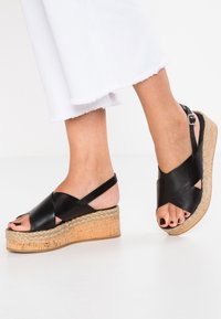 Černé kožené klínové sandály s křížovými páskami, střední částí obalenou jutou a texturovanou korkovou podešví. Design s otevřenou špičkou a nastavitelným páskem kolem kotníku.