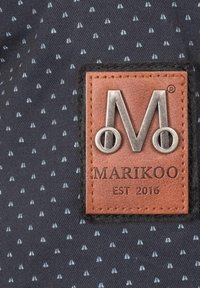 Mornarska tkanina z majhnimi svetlo modrimi trikotnimi vzorci, ki vsebuje pravokotno rjavo usnjeno oznako s srebrnim embosiranim logotipom "M" in besedilom "MARIKOO EST 2016."