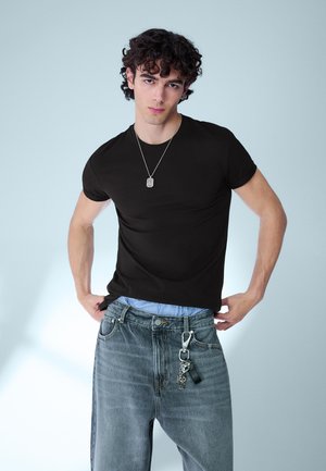 Mladý muž s kučeravými vlasmi, oblečený v čiernom tričku, sivých džínsoch s kľúčenkou a striebornom náhrdelníku, stojaci pred svetlým pozadím.