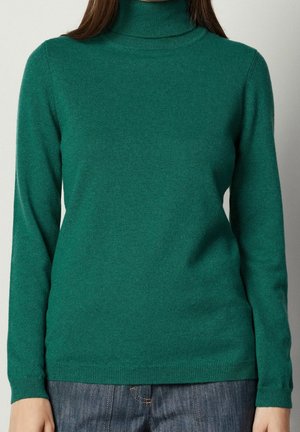 Personne portant un pull à col roulé vert cintré et un jean en denim bleu, visible du cou jusqu'aux hanches sur un fond uni.