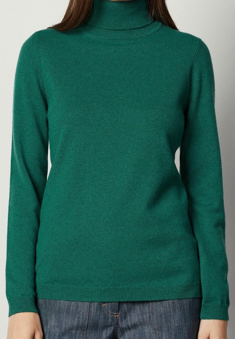 Personne portant un pull à col roulé vert cintré et un jean en denim bleu, visible du cou jusqu'aux hanches sur un fond uni.