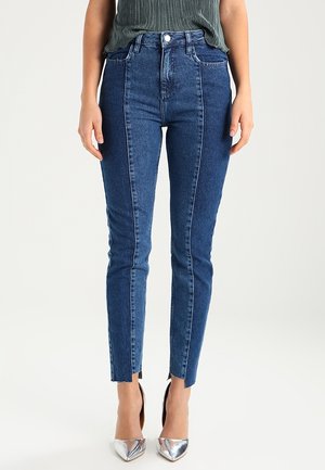 Kobieta nosząca dżinsy typu skinny z wysokim stanem w dwóch odcieniach niebieskiego z przeszyciem z przodu i rozcięciami na przodzie nogawek, zestawione z srebrnymi szpiczastymi szpilkami.