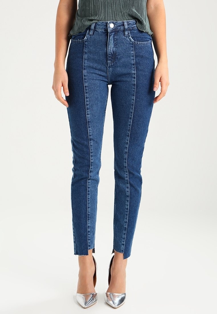 Femme portant un jean skinny bleu bicolore taille haute avec une couture à l'avant et des fentes à l'ourlet avant, associé à des escarpins pointus argentés.