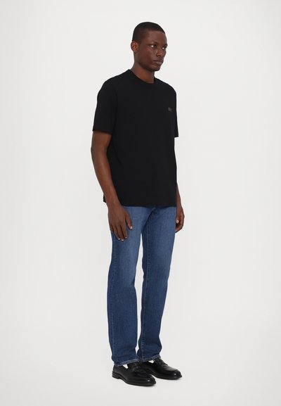 T-shirt en coton noir à manches courtes et col rond, arborant un petit logo multicolore sur la poitrine gauche ; associé à un jean bleu et des chaussures noires.