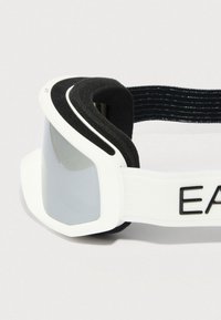 Λευκά γυαλιά σκι με αντανακλαστικούς φακούς, μαύρο αφρώδες υλικό εσωτερικά και ρυθμιζόμενο λευκό λουράκι με μαύρα γράμματα.