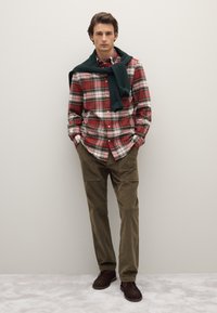 Rot-grün kariertes Hemd, olivfarbene Cordhosen, dunkelbraune Schuhe, mit einem dunkelgrünen Pullover, der über die Schultern gelegt ist. Einfacher, lässiger Stil.