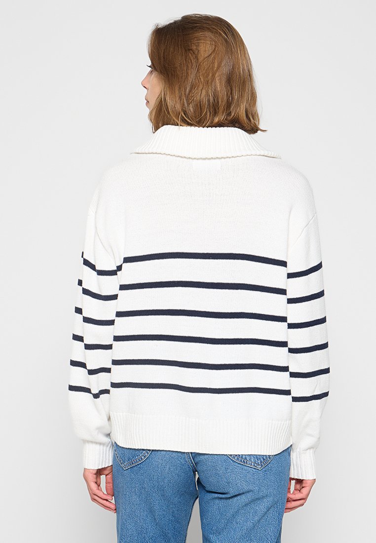 Persona con cabello corto castaño que lleva un suéter de punto blanco con rayas negras horizontales, combinado con jeans azules, de espaldas a la cámara.