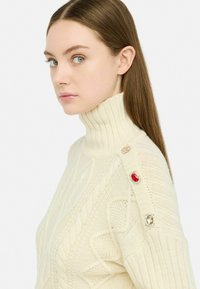 Maglione a collo alto in maglia crema con motivi a treccia, caratterizzato da accenti in gemme rosse e trasparenti sulla spalla. Texture liscia, forma aderente.