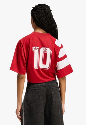 Persona con capelli intrecciati che indossa una maglia sportiva rossa con strisce bianche e numero 10 sul retro, abbinata a jeans neri a vita alta.