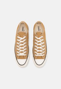 Zapatillas de lona marrón con cordones blancos, puntera de goma, ribete negro y suela plana. Logo visible en la lengüeta.