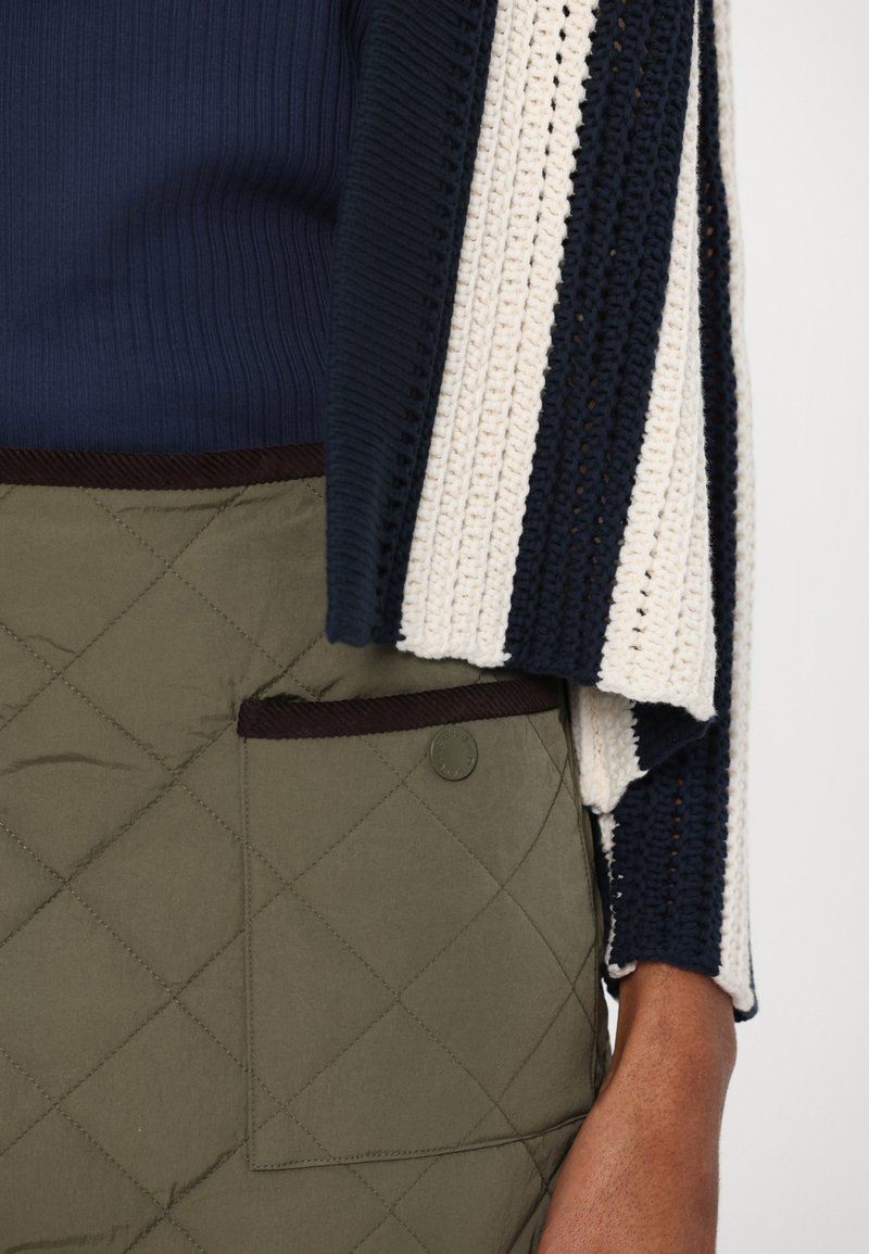 Top de canalé azul marino debajo de un cárdigan de ganchillo a rayas crema y azul marino; pantalones de oliva acolchados con un bolsillo de parche y ribetes oscuros.