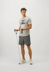 Pilka trumpomis rankovėmis marškinėliai su juodu logotipu, derinami su tamsiomis šortais, laikančiais pasipriešinimo juostas, dėvintys šviesių spalvų sportinius batelius.