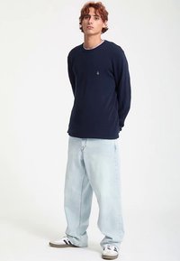 Marineblauw shirt met lange mouwen en geribbelde halslijn, gecombineerd met lichtblauwe jeans met wijde pijpen en witte sneakers met zwarte strepen.