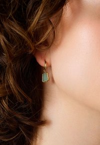 Guld hoop örhängen med en texturerad, grön sten med en organisk form hängande från ringen, förhöjd av intrikata detaljer.