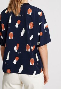 Navy blue oversized shirt featuring abstract face patterns in orange, cream, and white. Short sleeves and a relaxed fit.