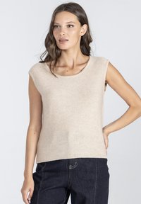 Beżowy, dziany top na ramiączkach z okrągłym dekoltem i krótkimi rękawami, z gładką fakturą i prążkowanym wykończeniem, w zestawie z ciemnoniebieskimi dżinsami.
