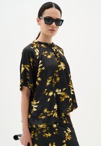 Blusa nera con motivi floreali gialli, tessuto leggero, scollatura arricciata, maniche corte. Indossata con occhiali da sole scuri, mentre tiene un manico nero.