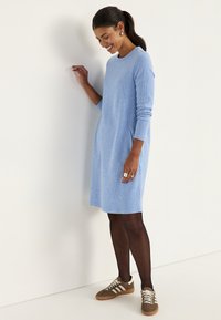 Hellblaues Strickkleid mit langen Ärmeln, Rundhalsausschnitt, Seitentaschen und lockerem Schnitt, kombiniert mit braunen Sneakers und dunklen Strumpfhosen.