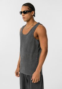 Donkergrijs mouwloos tanktop gemaakt van ribbelstof met een brede halslijn. Heeft rauwe randen en een losse pasvorm, gedragen met bijpassende broeken.