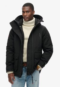 Chaqueta de invierno negra con un alto cuello, múltiples bolsillos frontales y cierre de cremallera. Lleva puesta sobre un suéter beige y unos vaqueros desgastados.