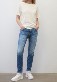 Světle béžové tričko s krátkým rukávem, modré úzké džíny s bledými nášivkami a bílé tenisky s šedým akcentem. Neformální outfit na modelu.