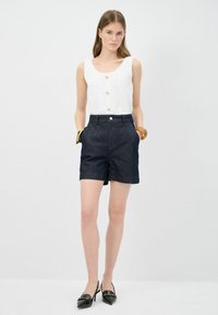 Vit texturerad linne med gyllene knappar, kombinerad med mörkblå knälånga shorts med sidofickor och en åtsittande midja. Svarta klackskor.