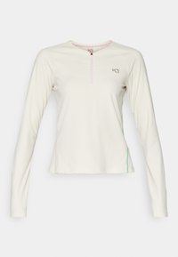 Hladký krémový top s dlouhým rukávem a půl zipem. Obsahuje ribbed texturu, růžový zip a zelený akcentní šev po straně. Kulatý výstřih.