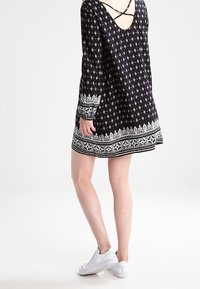 Vestido negro con patrón blanco, mangas largas, escote en V con tirantes cruzados y un dobladillo decorativo. Combinado con zapatillas blancas.
